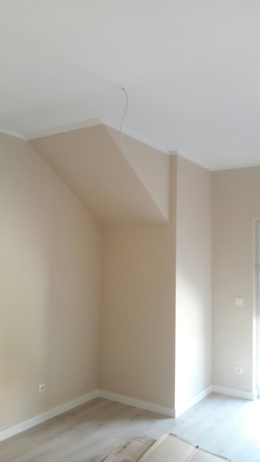 Pomalowane na beżowo wnętrze pokoju z widoczną instalacją elektryczną wystającą z sufitu, podłoga wyłożona panelami, ściany w jasnym odcieniu beżu.