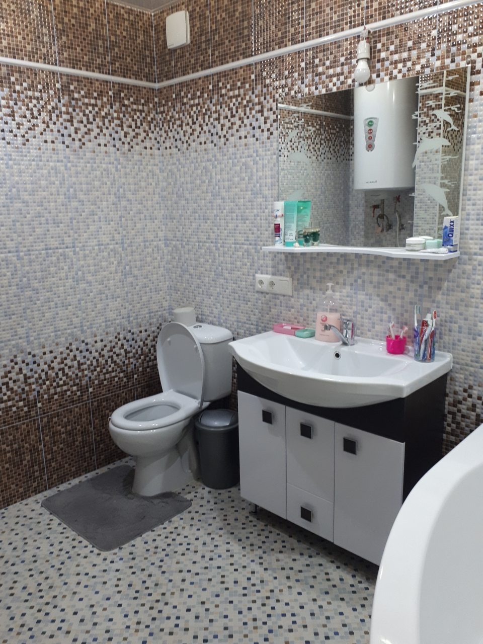 Ukończona łazienka z mozaikową glazurą, białą umywalką z szafką, toaletą i bojlerem widocznym w lustrze.