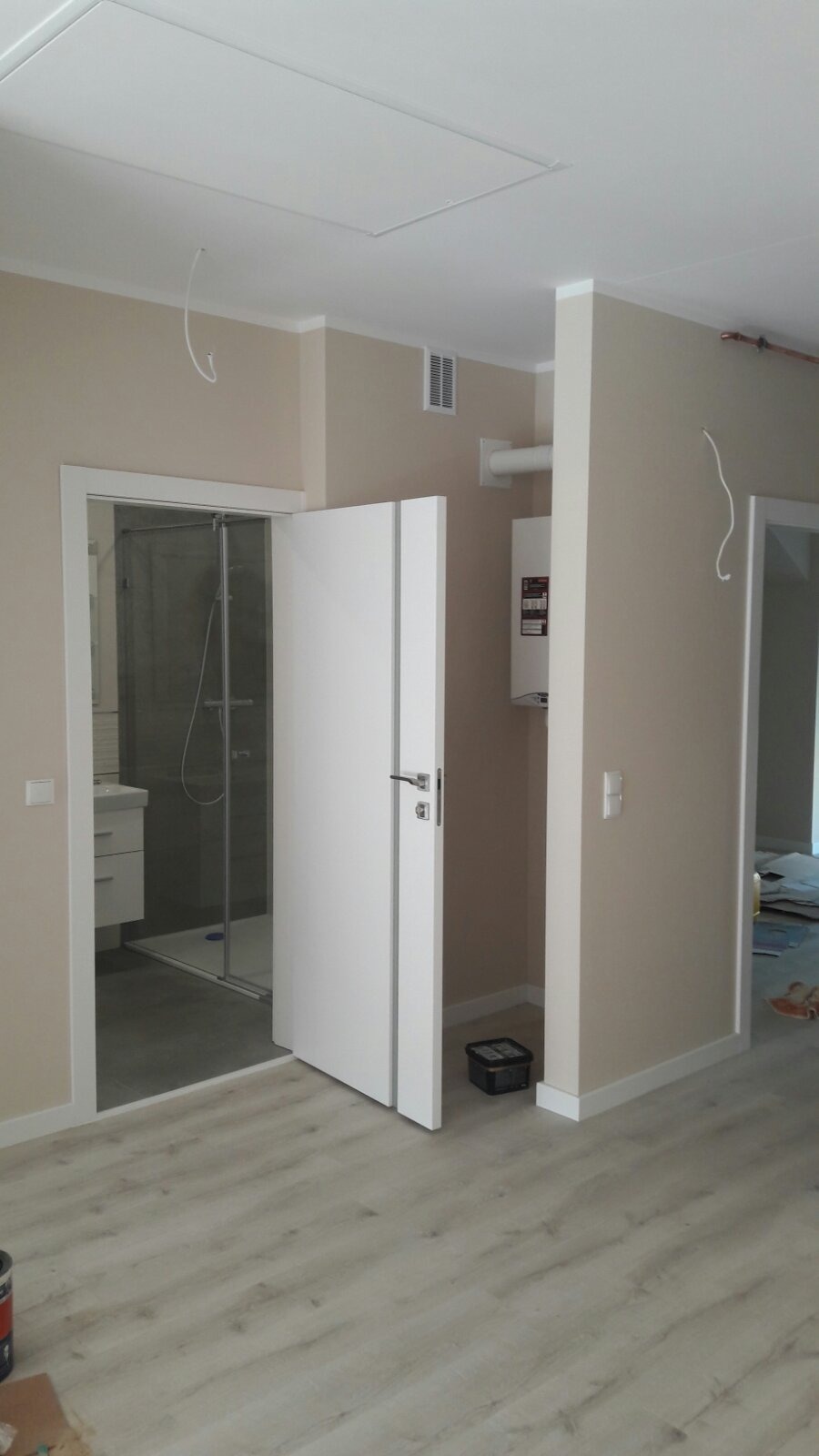 Wnętrze mieszkania w trakcie wykańczania, z widoczną otwartą białą łazienką z kabiną prysznicową, fragmentem podłogi z paneli, grzejnikiem na ścianie i elementami instalacji pod sufitem.