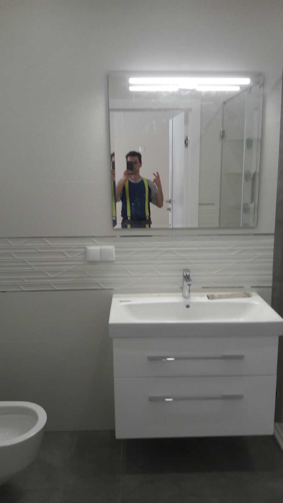 Wykończona łazienka z białą umywalką nablatową z szafką, widoczne dekoracyjne płytki na ścianie i odbicie glazurnika robiącego zdjęcie w lustrze.