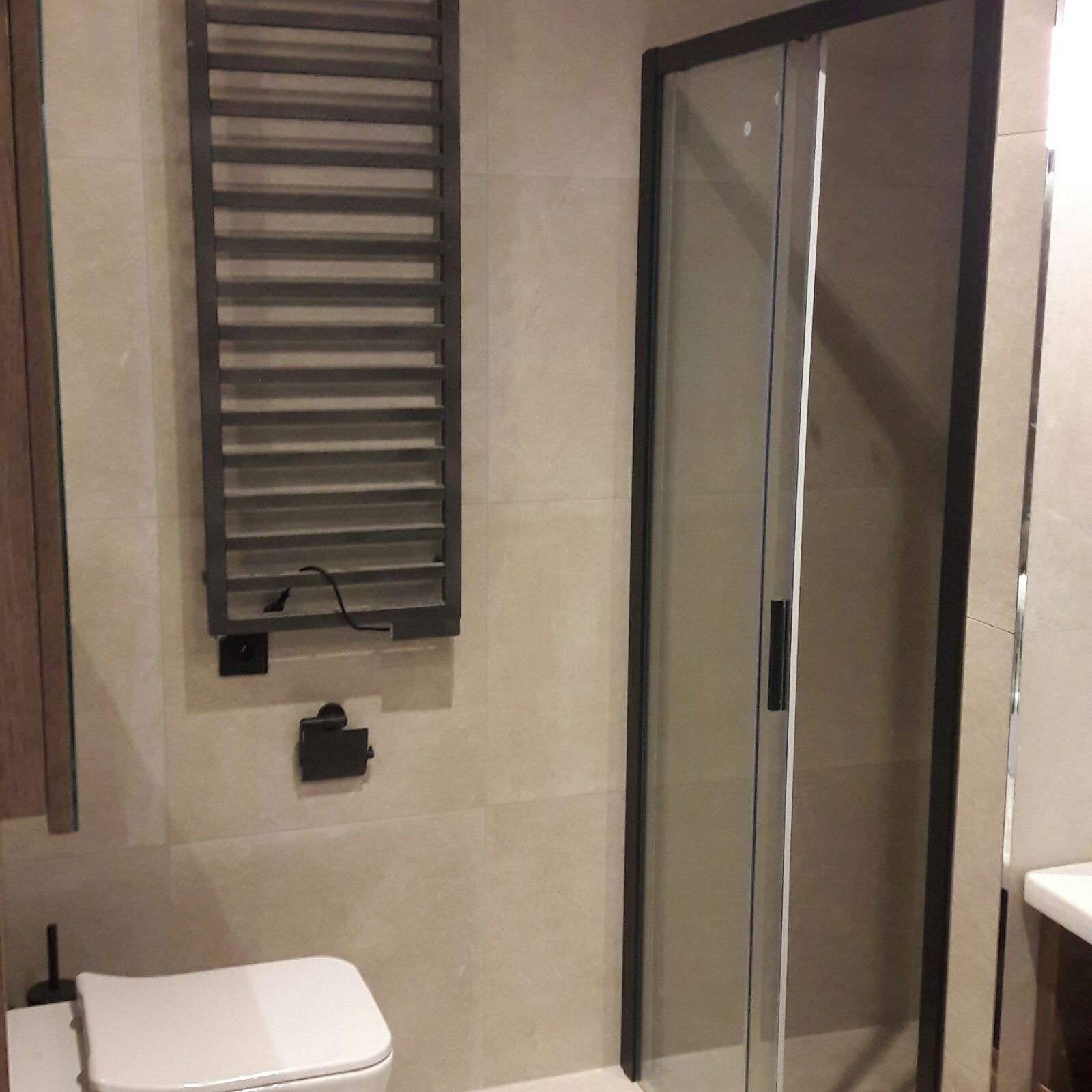 Nowoczesna łazienka z beżowymi płytkami, grzejnikiem drabinkowym w kolorze antracytowym, kabiną prysznicową z czarnymi profilami i podwieszaną toaletą.