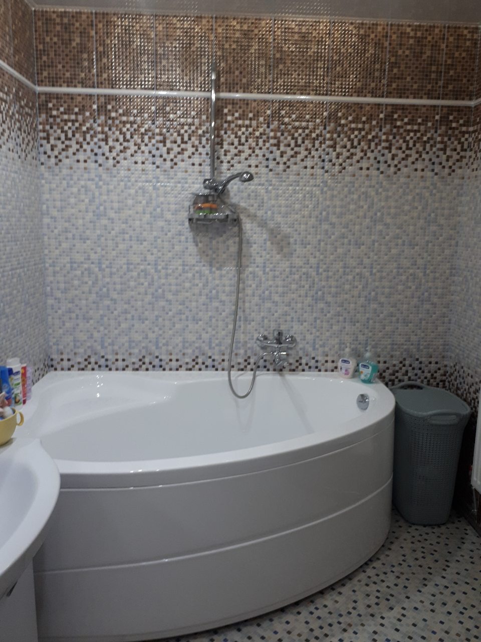 Biała wanna narożna z baterią i prysznicem na tle mozaiki w odcieniach brązu i błękitu, uzupełniona szarym koszem na pranie i kosmetykami.