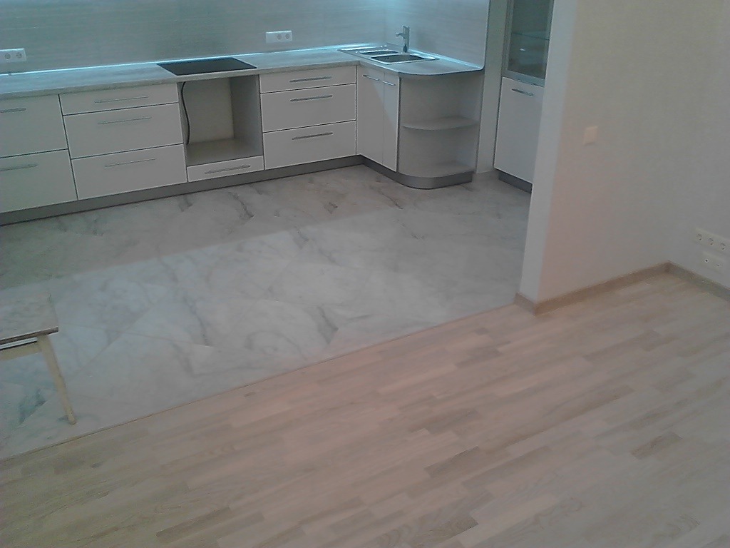 Nowoczesna biała kuchnia z marmurową podłogą i drewnianym parkietem w sąsiednim pomieszczeniu, widoczne gniazdka elektryczne na ścianie.