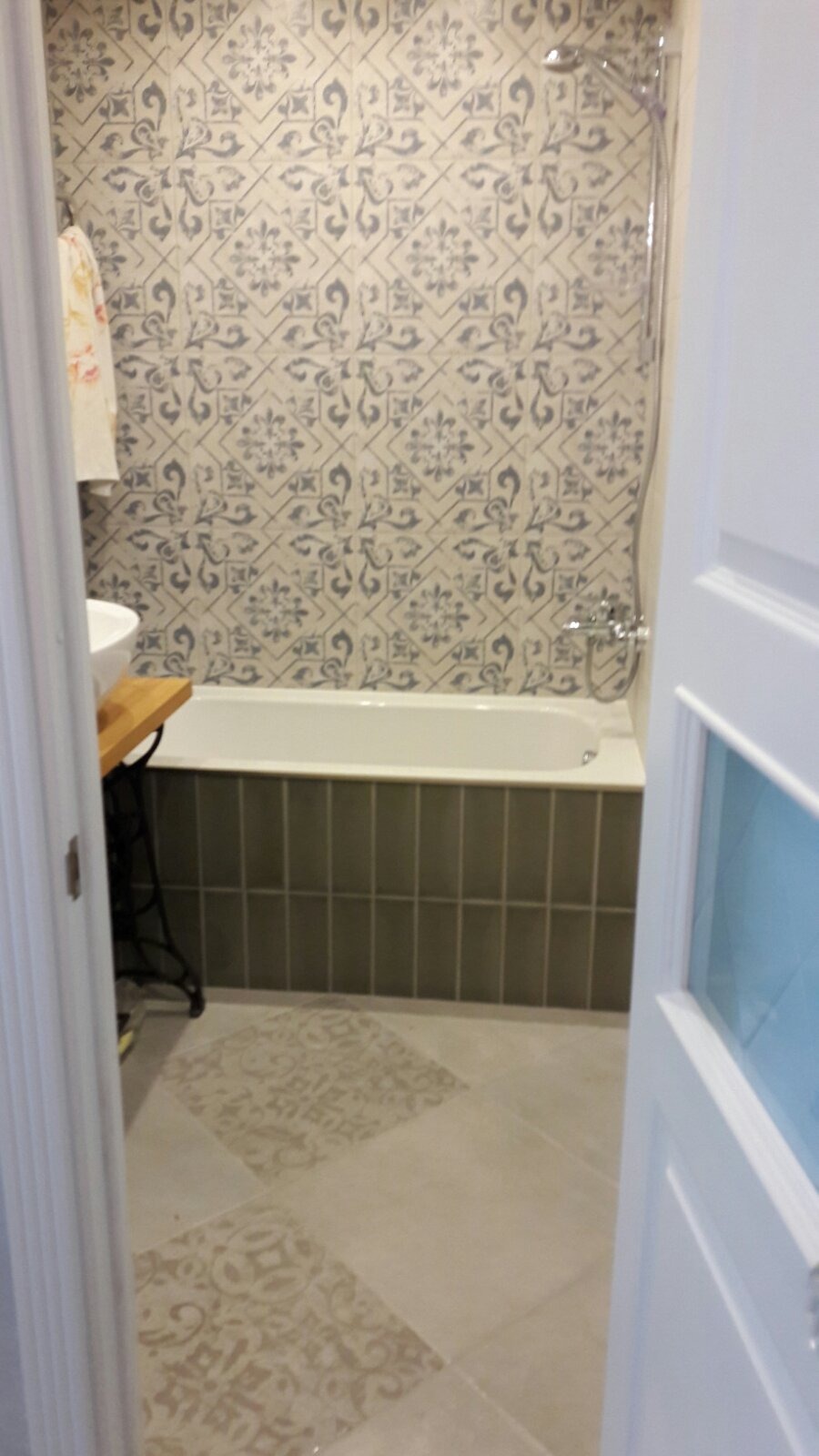 Wykończona łazienka z wanną obudowaną szarymi, pionowo ułożonymi płytkami, wzorzystą ścianą nad wanną, oraz wzorzystą podłogą, widoczna przez uchylone białe drzwi.