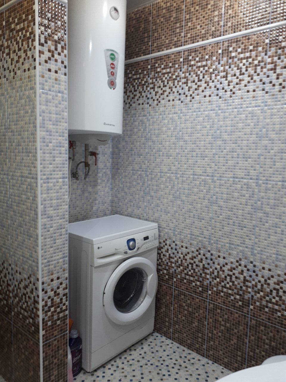 Łazienka po remoncie z mozaikowymi płytkami w odcieniach brązu i błękitu, z białą pralką i podgrzewaczem wody Ariston.