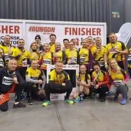 Grupa biegaczy w żółtych koszulkach z napisem 'Train Now' pozuje po ukończonym maratonie w Gdańsku, trzymając medale i numery startowe.