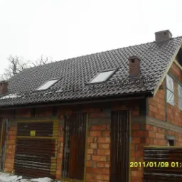 Budynek w trakcie budowy, z czerwonej cegły, z zamontowanym brązowym dachem z dachówkami, dwoma oknami dachowymi i kominami, okna na parterze zabezpieczone folią.