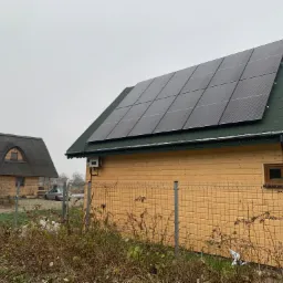 Panele słoneczne na dachu drewnianego domu z zieloną dachówką, w tle drugi dom z dachem krytym strzechą i metalowe ogrodzenie.