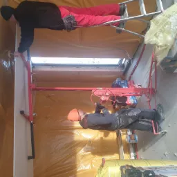 Dwóch pracowników montuje płytę kartonowo-gipsową na suficie poddasza, zabezpieczonym żółtą folią ochronną; jeden używa podnośnika, drugi stoi na drabinie, widoczne narzędzia i materiały budowlane.