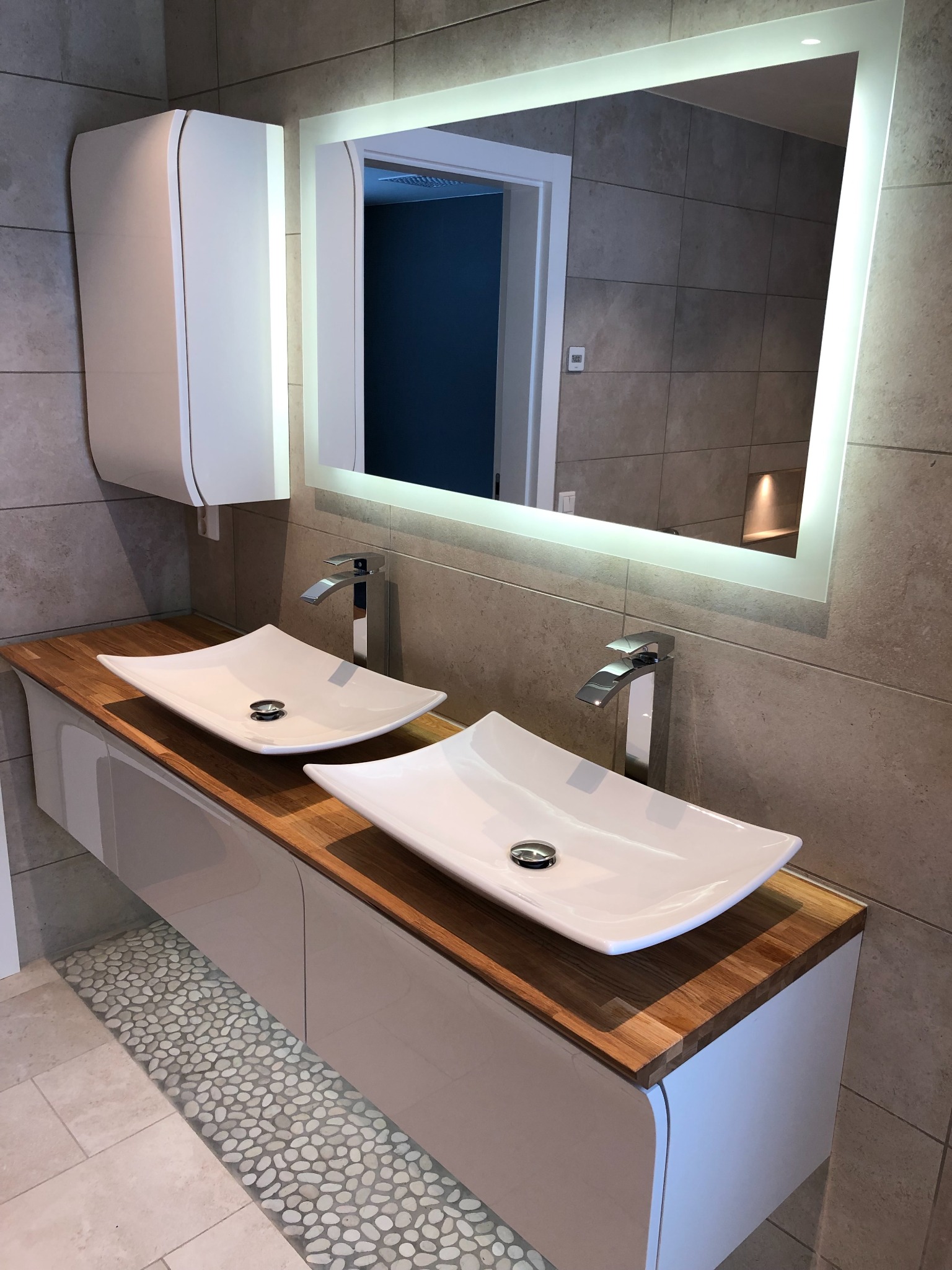 Nowoczesna łazienka z podwójną umywalką nablatową na drewnianym blacie, podświetlane lustro i biała szafka wisząca, płytki ceramiczne na ścianach i podłodze z dekoracyjnym pasem z kamyków.