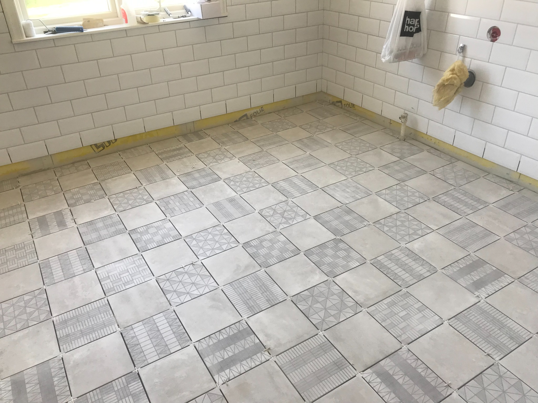 Układanie kwadratowych płytek podłogowych z geometrycznymi wzorami w jasnym pomieszczeniu, zabezpieczone folią przy ścianach z białymi, prostokątnymi płytkami.