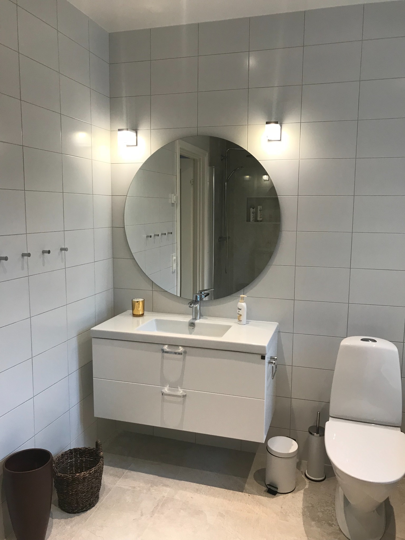 Nowoczesna łazienka z białymi płytkami, okrągłym lustrem podświetlonym dwoma lampami, wiszącą szafką z umywalką, toaletą i akcesoriami w stonowanej kolorystyce.