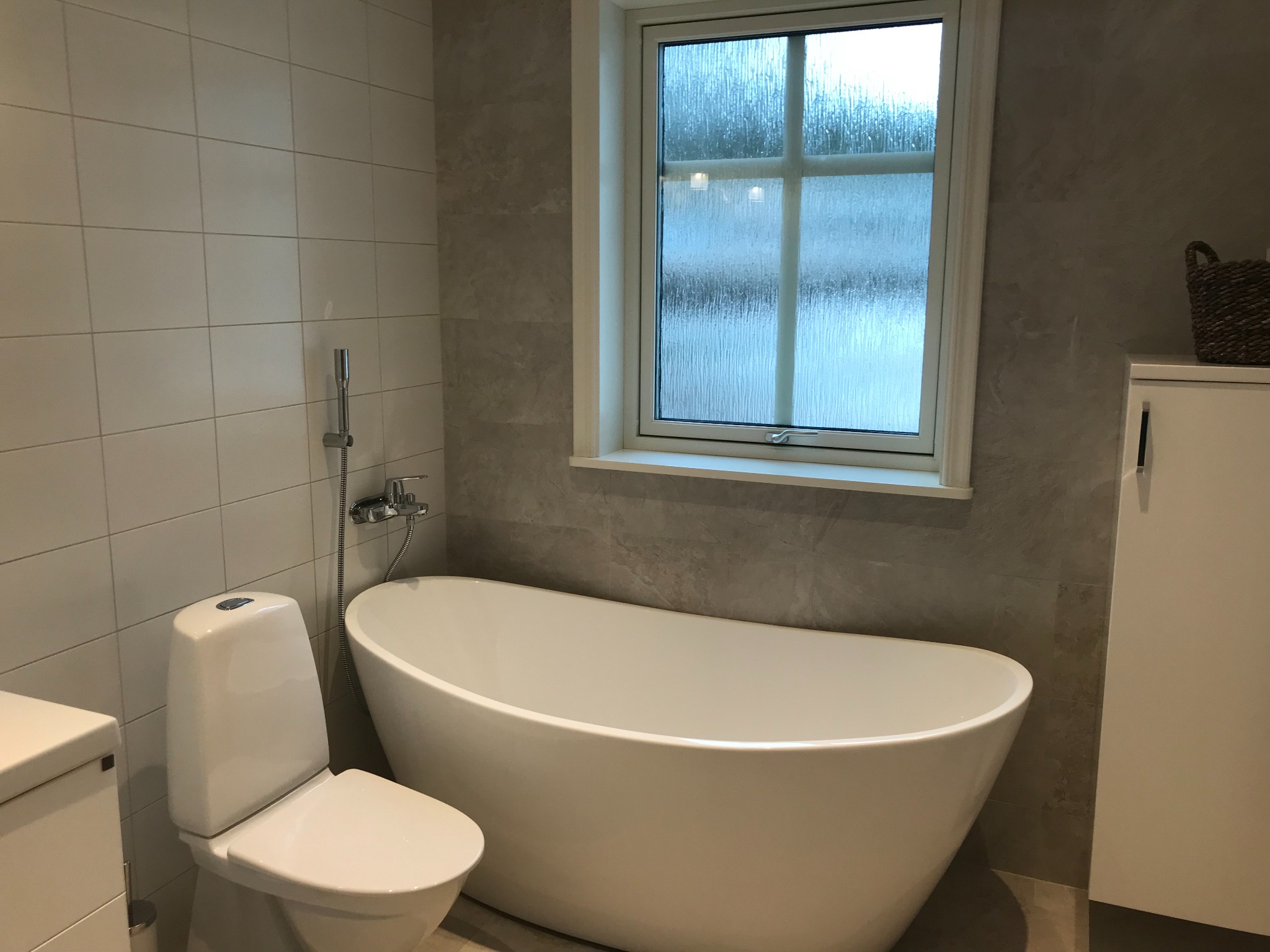 Nowoczesna łazienka z wolnostojącą wanną, toaletą, szafką pod umywalkę i oknem z teksturowaną szybą, wykończona płytkami w odcieniach szarości.