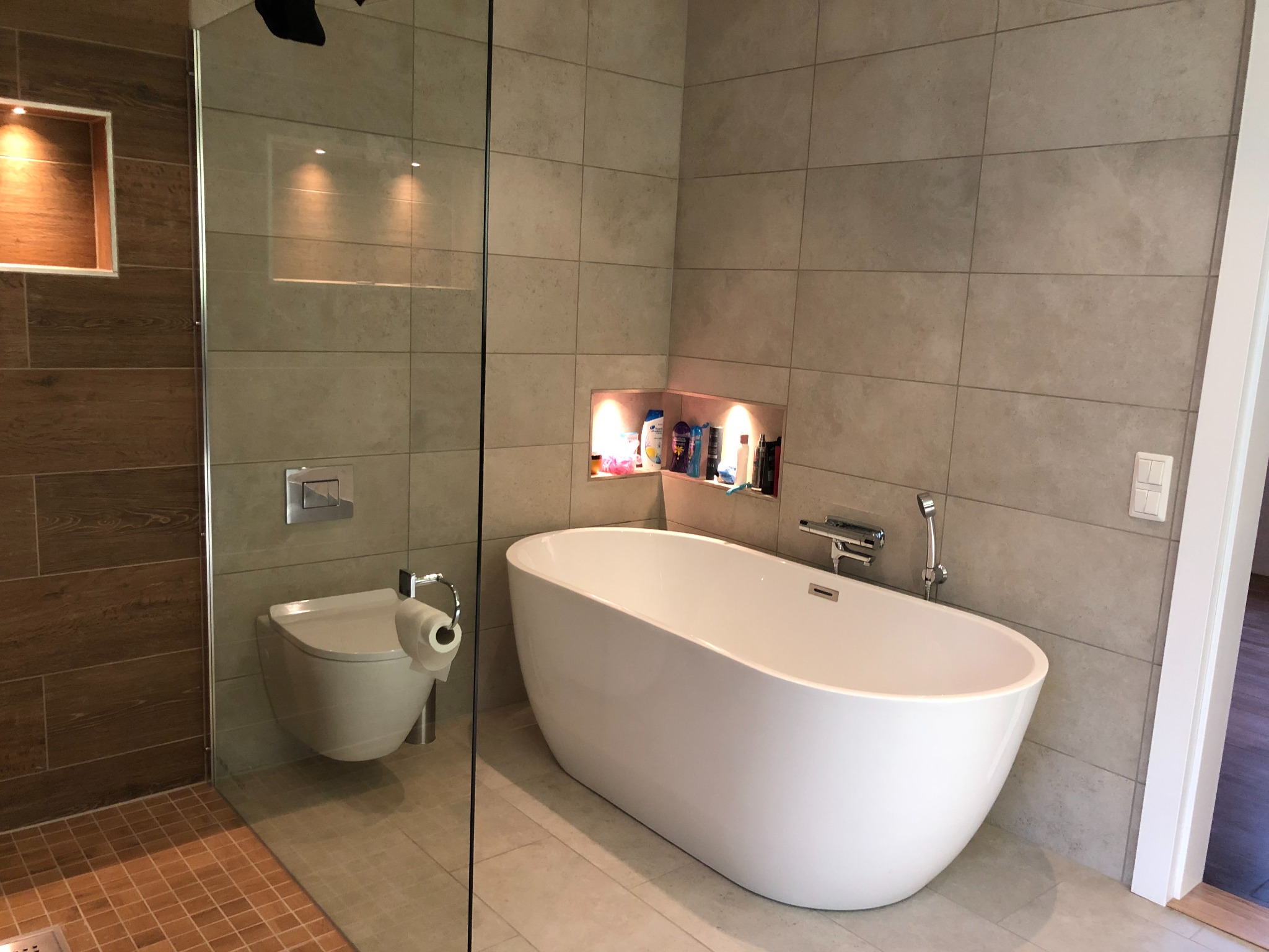 Nowoczesna łazienka z wolnostojącą wanną, podwieszaną toaletą i szklaną kabiną prysznicową, wykończona płytkami w odcieniach beżu i brązu, z podświetlanymi niszami na kosmetyki.