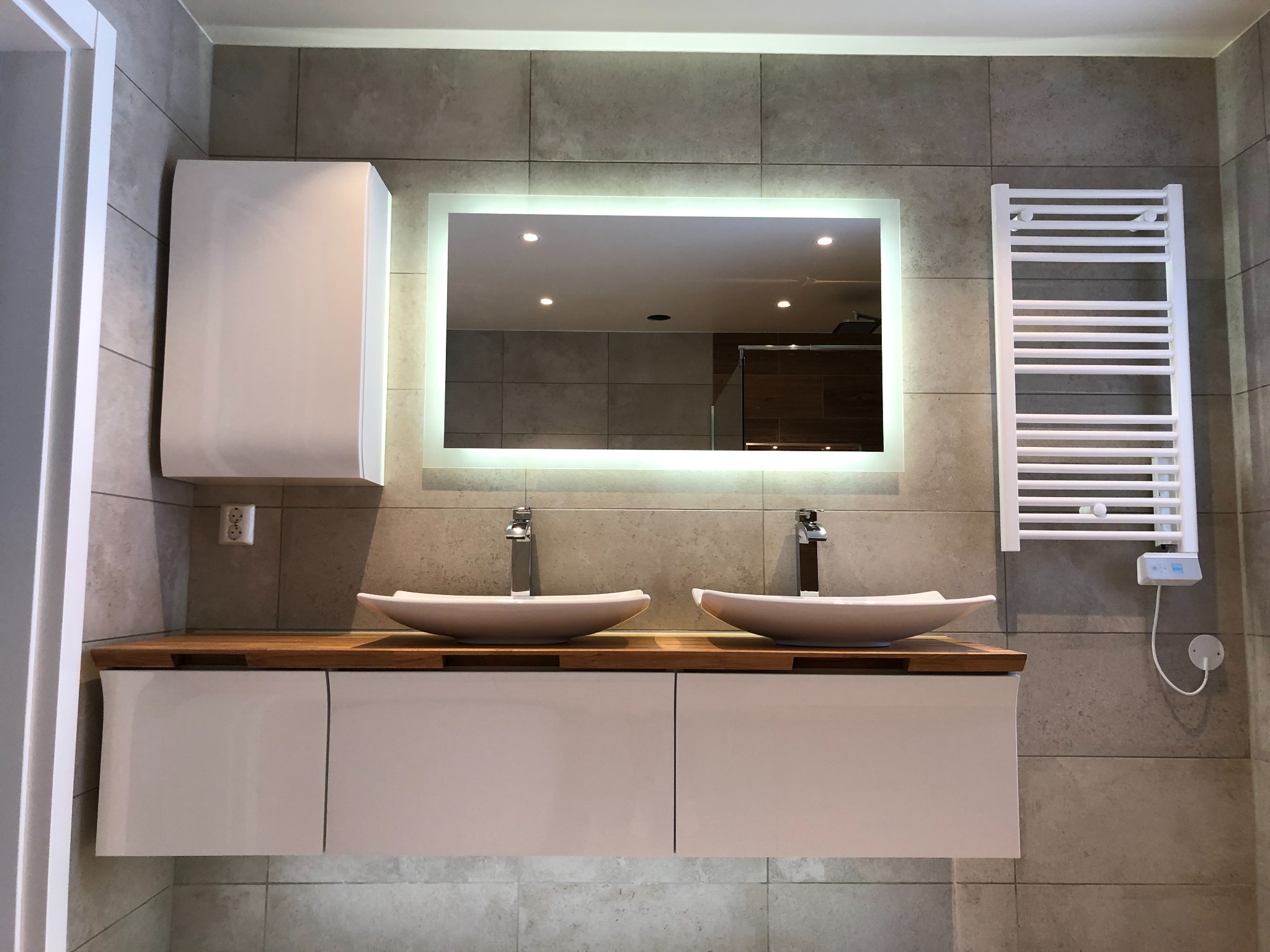 Nowoczesna łazienka z podwójną umywalką w kształcie łódki, drewnianym blatem, białymi szafkami wiszącymi, podświetlanym lustrem i grzejnikiem drabinkowym na szarych płytkach.