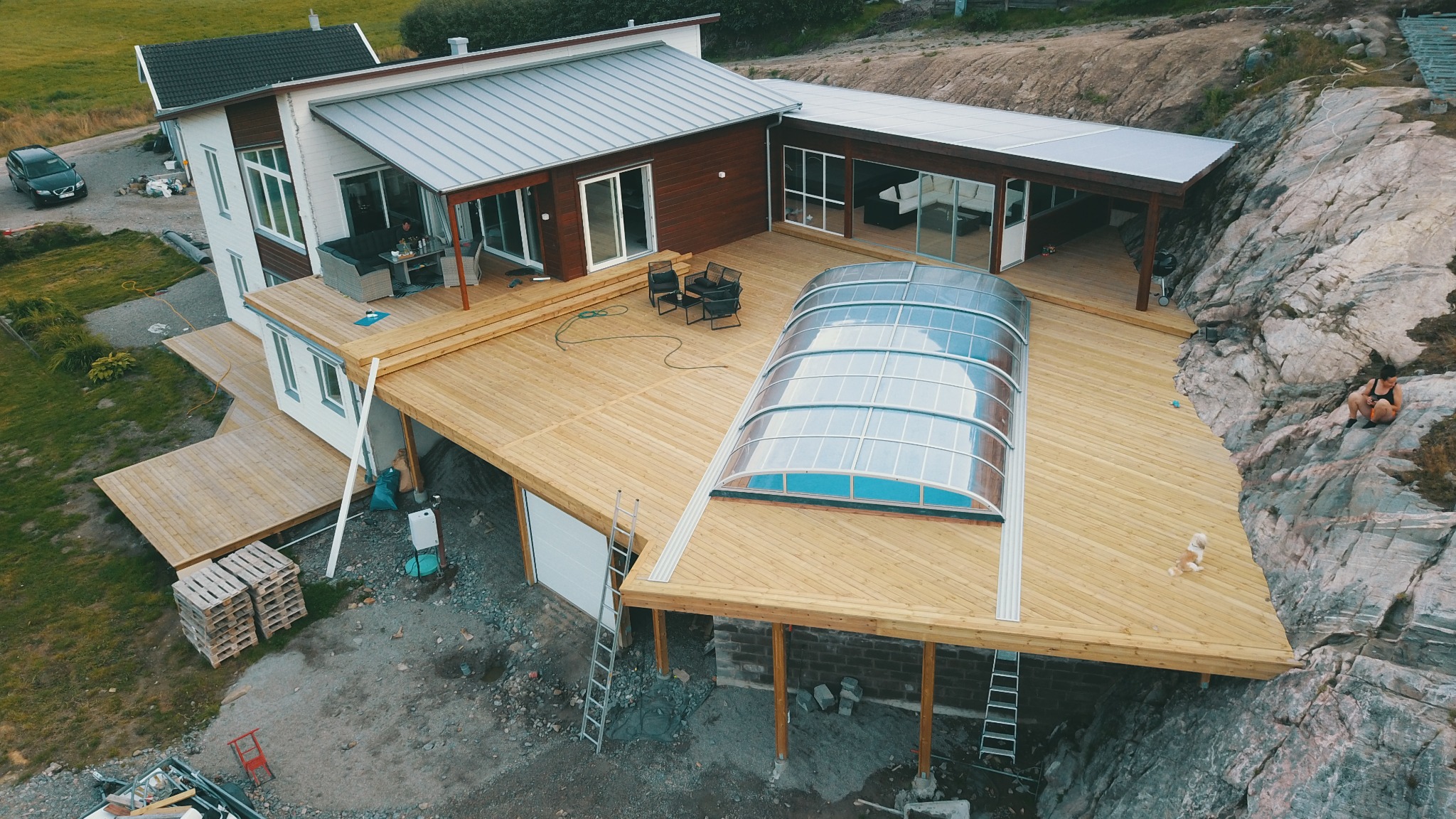 Nowoczesny dom z rozległym drewnianym tarasem na skalistym terenie, z zadaszeniem basenowym i dodatkowym zadaszonym patio, widok z góry.