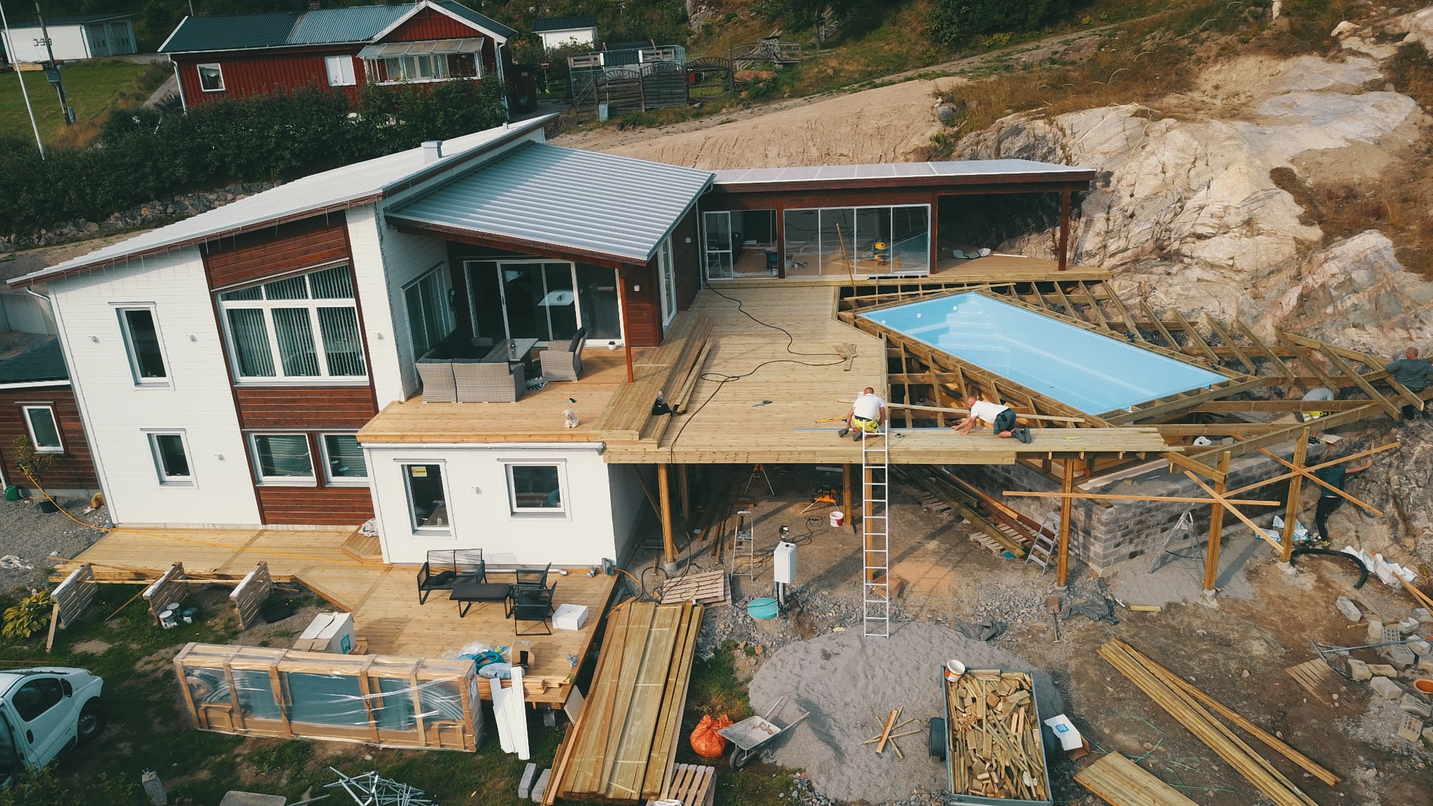 Widok z góry na budowę domu z drewnianym tarasem i basenem na zboczu skalnym, dwóch robotników przy pracy, materiały budowlane porozrzucane wokół.