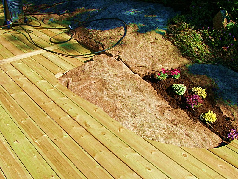 Drewniany taras z jasnych desek, wkomponowany w naturalny skalniak z kolorowymi kwiatami, widoczny wąż ciśnieniowy i myjka.