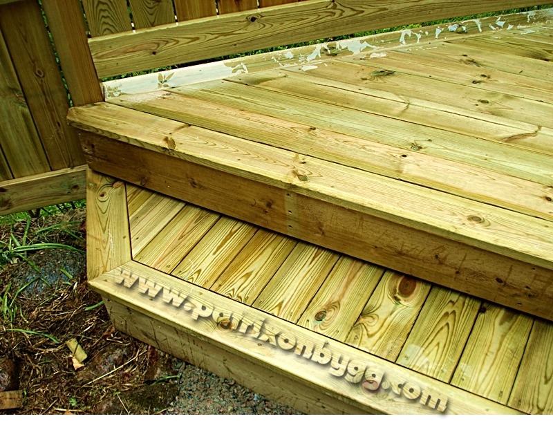 Drewniane schody tarasowe z widocznym usłojeniem i fragmentem ogrodzenia, świeżo po impregnacji, z widocznym adresem strony internetowej na dolnym stopniu.