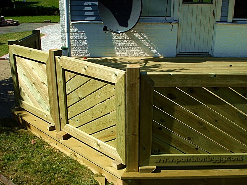 Drewniana balustrada tarasu z ukośnymi deskami, widok na elewację budynku z białą cegłą i anteną satelitarną.