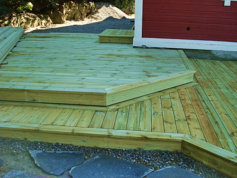 Nowo wybudowany drewniany taras przylegający do czerwonego budynku z białym obramowaniem, widoczne schodki i wykończenie z kamieni ozdobnych.