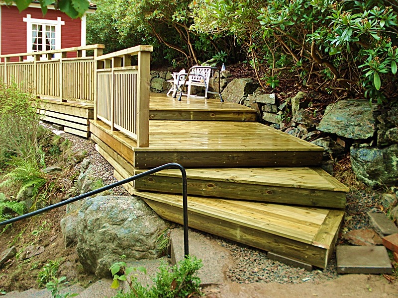 Drewniany taras z balustradą i schodami na nierównym terenie, z widokiem na ogród i elementy skalne.