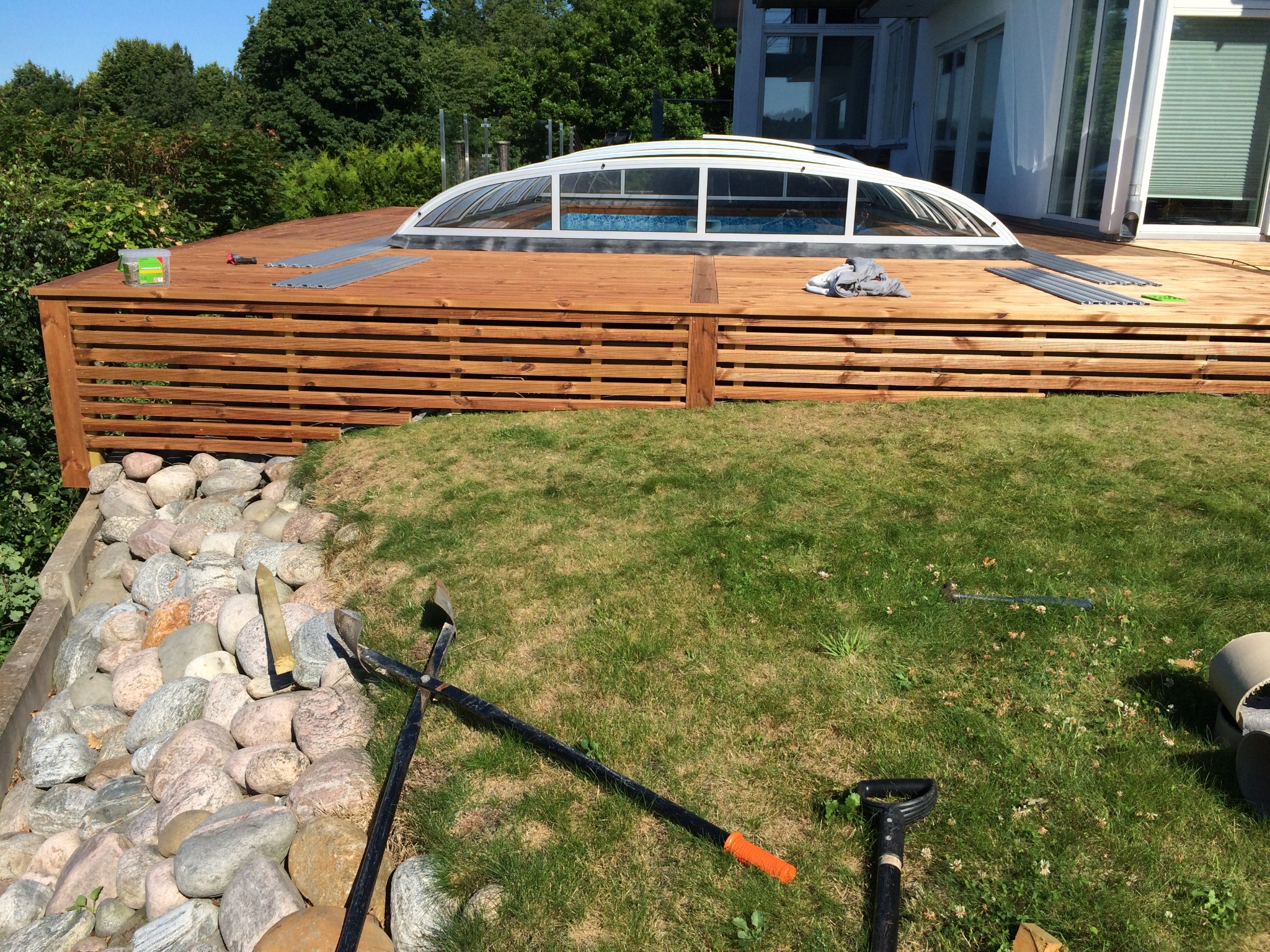 Drewniany taras z zadaszeniem basenu w trakcie budowy, widoczne narzędzia ogrodnicze i materiały budowlane na trawie.
