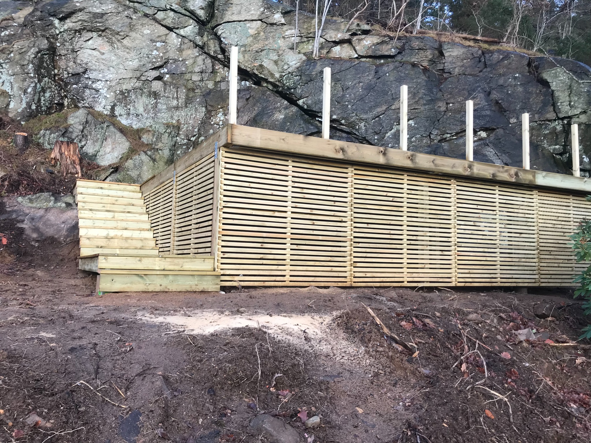 Drewniana konstrukcja z poziomych listew, prawdopodobnie taras lub podest, z drewnianymi schodami, usytuowana na nierównym terenie u podnóża skalnej ściany.