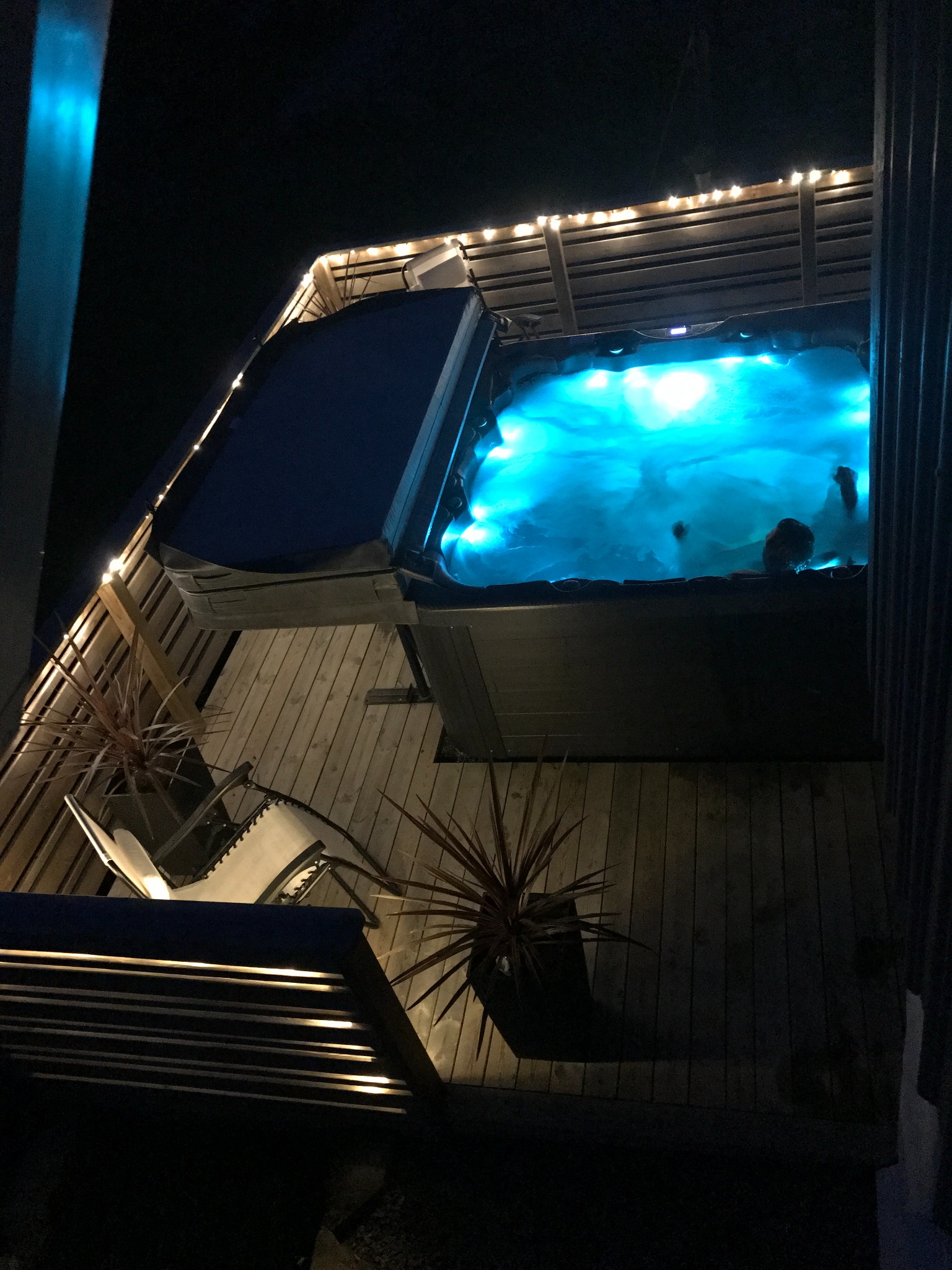 Zewnętrzne jacuzzi z podświetleniem LED w drewnianej altance, częściowo otwarte, widoczne osoby w wodzie, obok leżaki i rośliny doniczkowe, nocne oświetlenie.