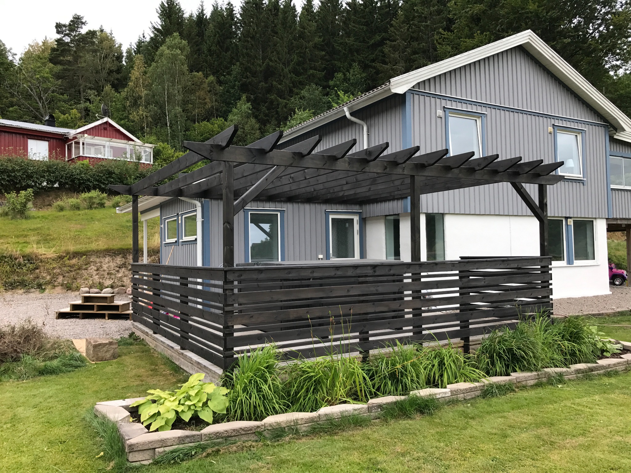 Szary dom z drewnianą elewacją, ciemną pergolą nad tarasem i starannie zagospodarowanym ogrodem z niskim kamiennym murkiem.