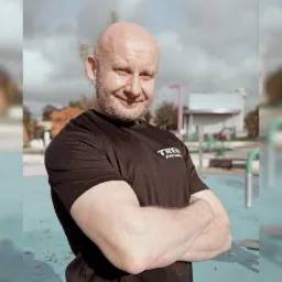 Uśmiechnięty, łysy mężczyzna z brodą, w czarnej koszulce z napisem na tle placu zabaw na zewnątrz.