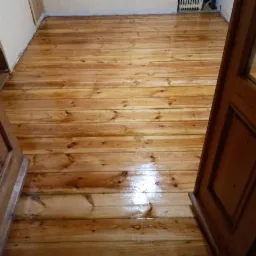 Świeżo polakierowana podłoga z desek, widoczne odbicia światła, drewniane drzwi w tle.