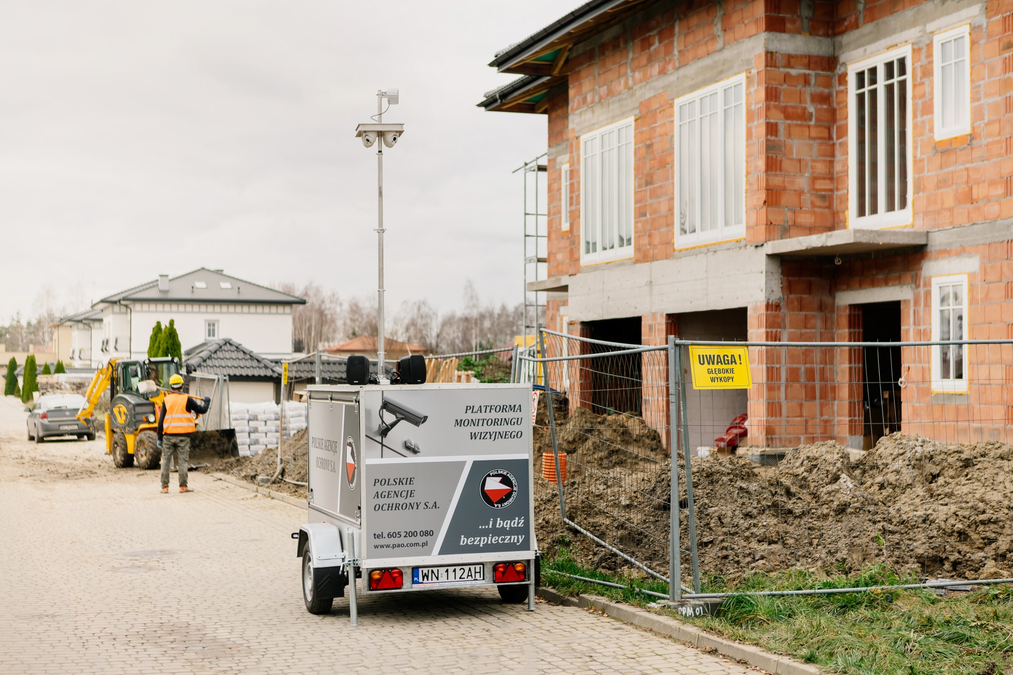 Mobilna platforma monitoringu wizyjnego z kamerami na przyczepie, z logo agencji ochrony, ustawiona na terenie budowy domu jednorodzinnego, w tle pracujący operator koparki.