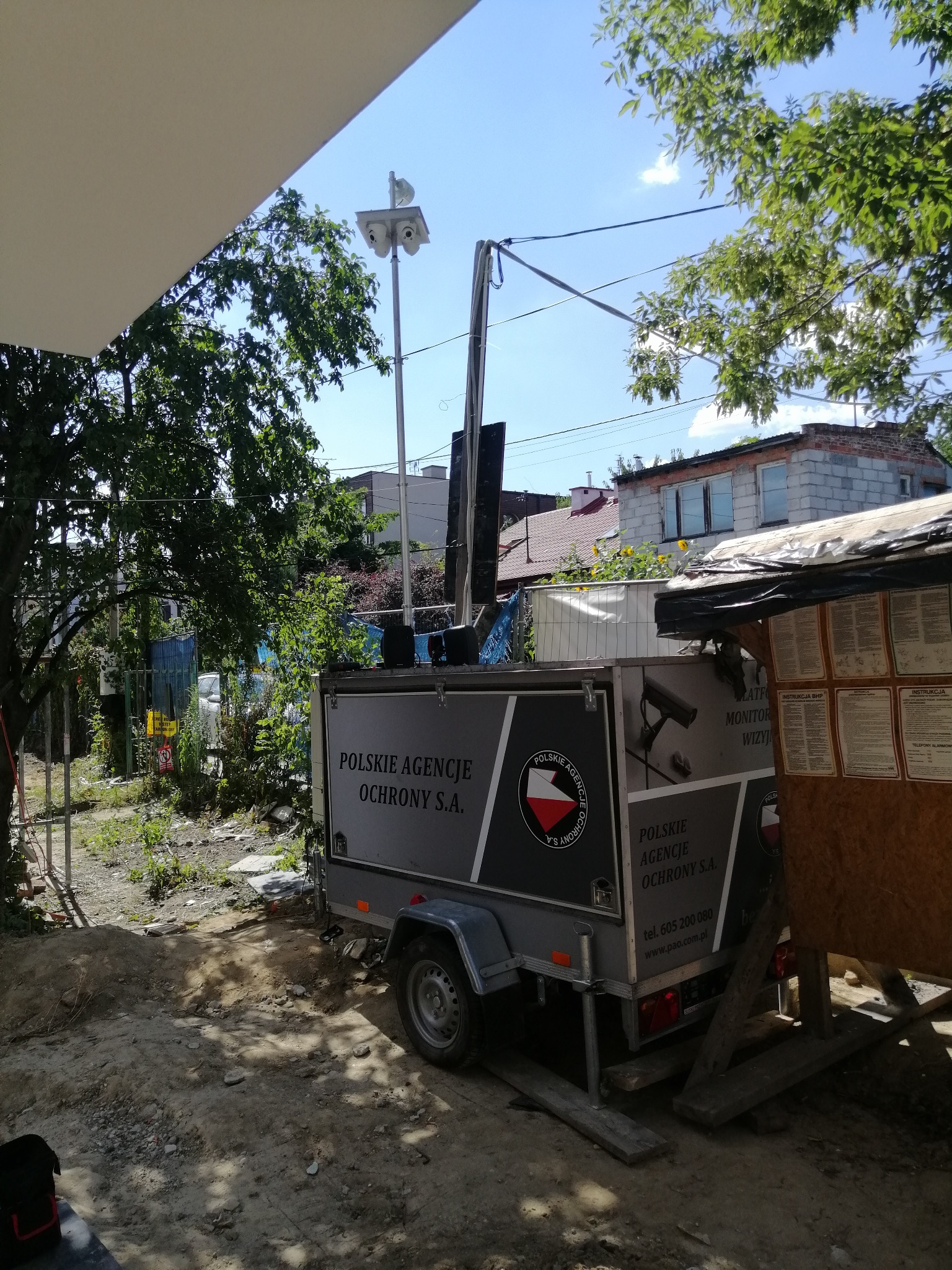Mobilny system monitoringu wizyjnego na przyczepie z logo firmy, ustawiony na placu budowy z widokiem na budynek mieszkalny w tle.