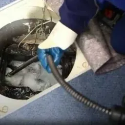 Pracownik w niebieskich rękawicach udrażnia zatkany odpływ podłogowy za pomocą specjalistycznego węża i urządzenia mechanicznego, generując pianę.