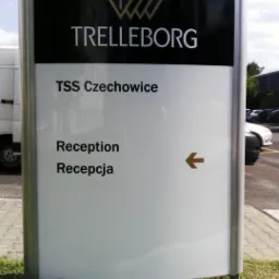 Pylon reklamowy z logo Trelleborg i napisem TSS Czechowice, wskazujący strzałką na recepcję w dwóch językach.
