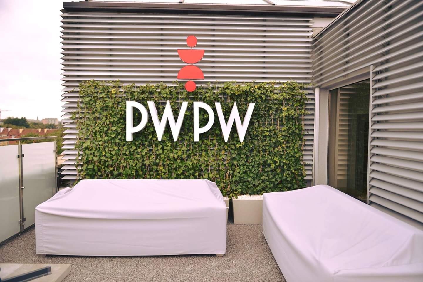 Zewnętrzna ściana biura z logo firmy PWPW, umieszczonym na tle z zielonych pnączy, z widocznymi meblami ogrodowymi przykrytymi białym materiałem.