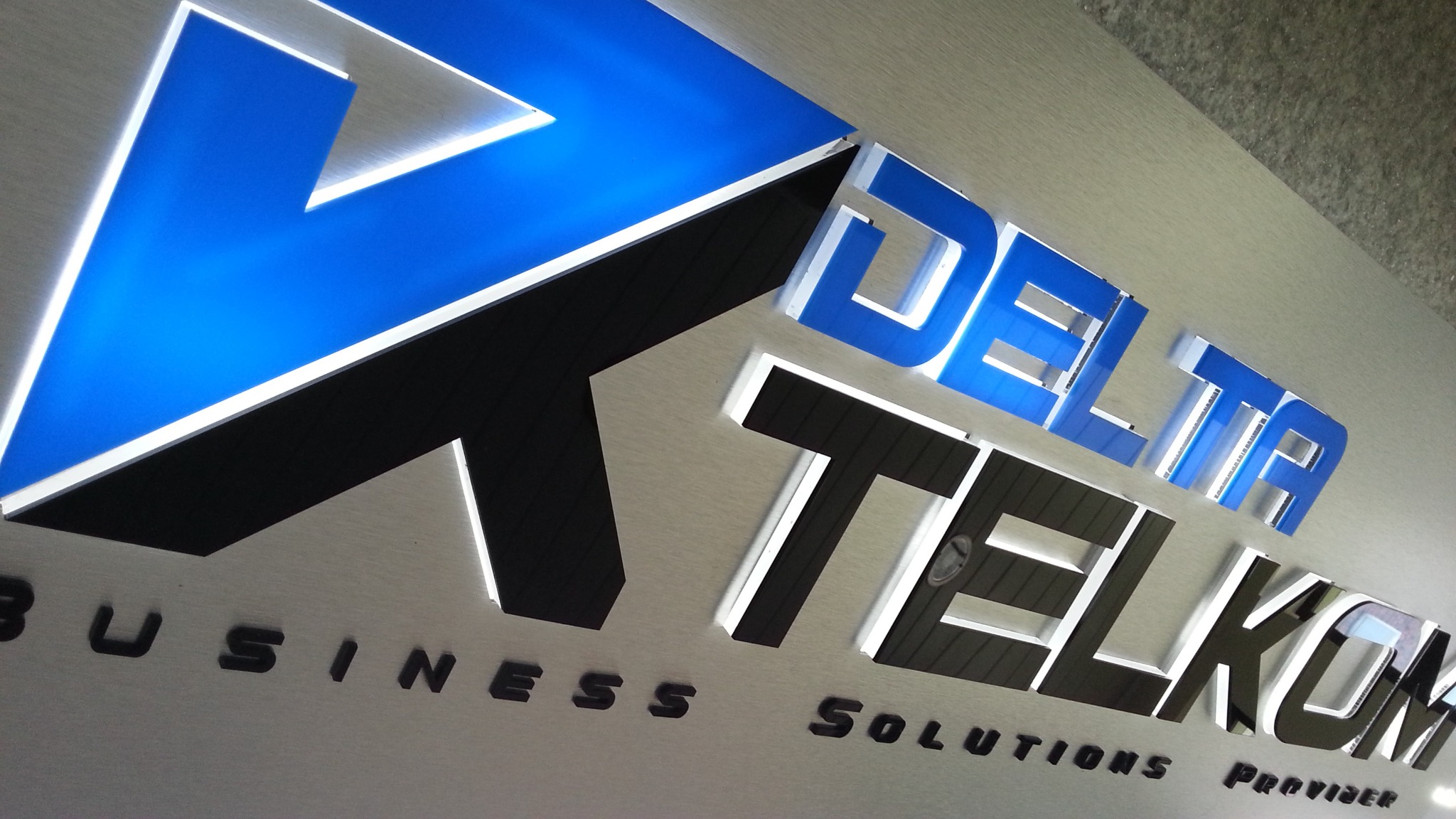 Podświetlane logo firmy Delta Telkom na srebrnym tle z napisem Business Solutions Provider w dolnej części, wykonane z czarnych i niebieskich elementów.
