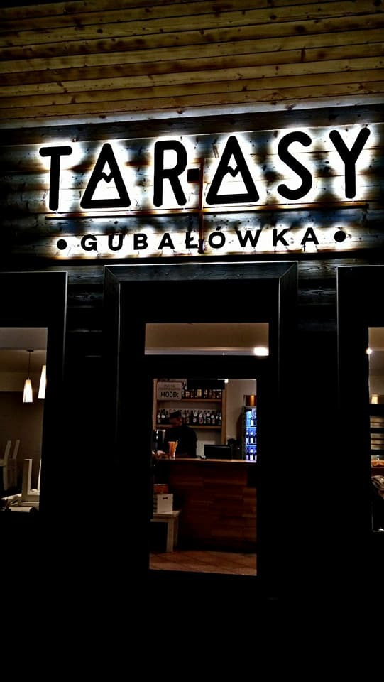 Podświetlany szyld 'Tarasy Gubałówka' na drewnianej elewacji, widok z zewnątrz na wnętrze lokalu z barem i obsługą.