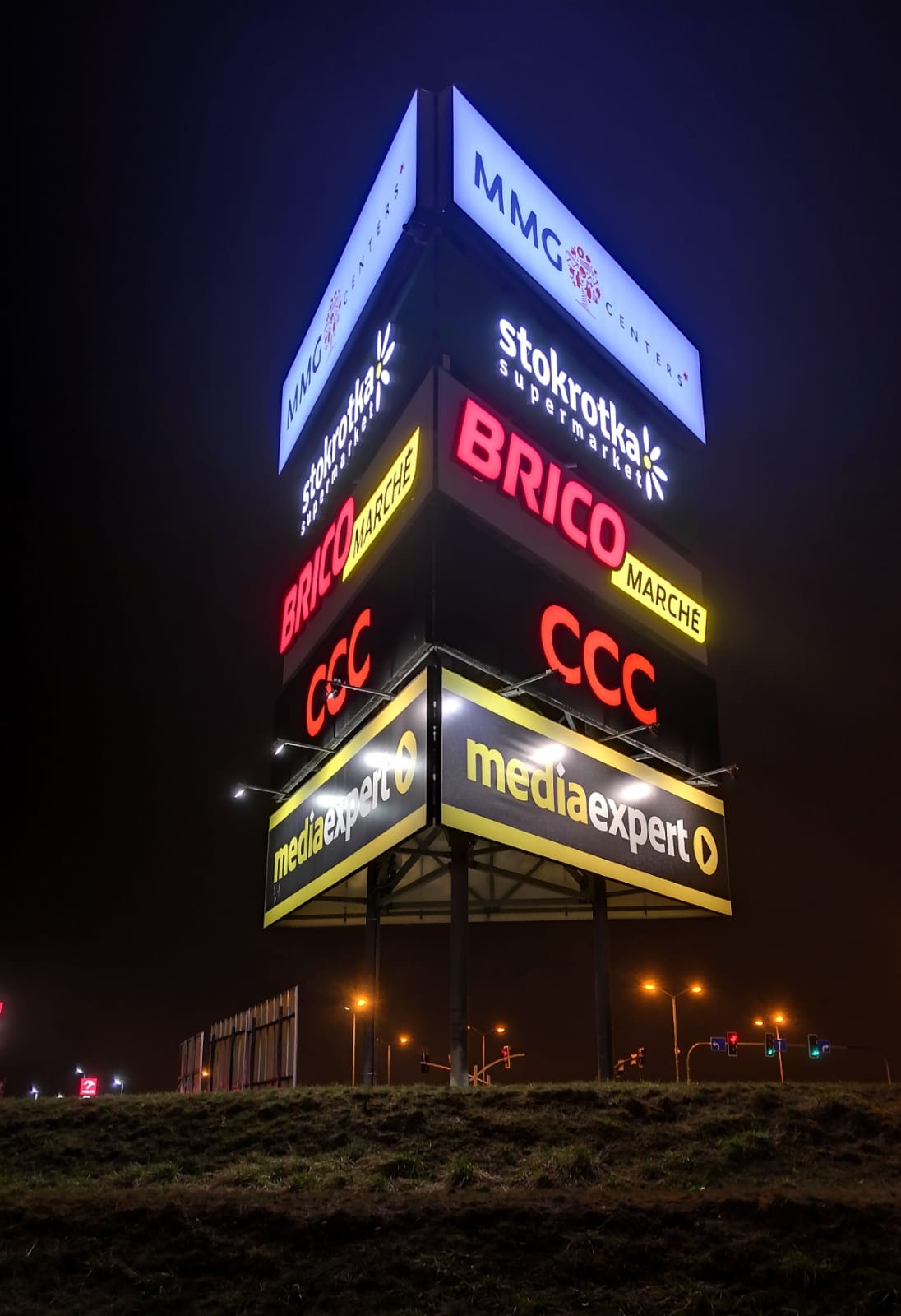 Wielkopowierzchniowa, podświetlana reklama zewnętrzna z logotypami różnych firm (MMG Centers, Stokrotka, Brico Marche, CCC, Media Expert) w Warszawie, widoczna nocą z oddali.