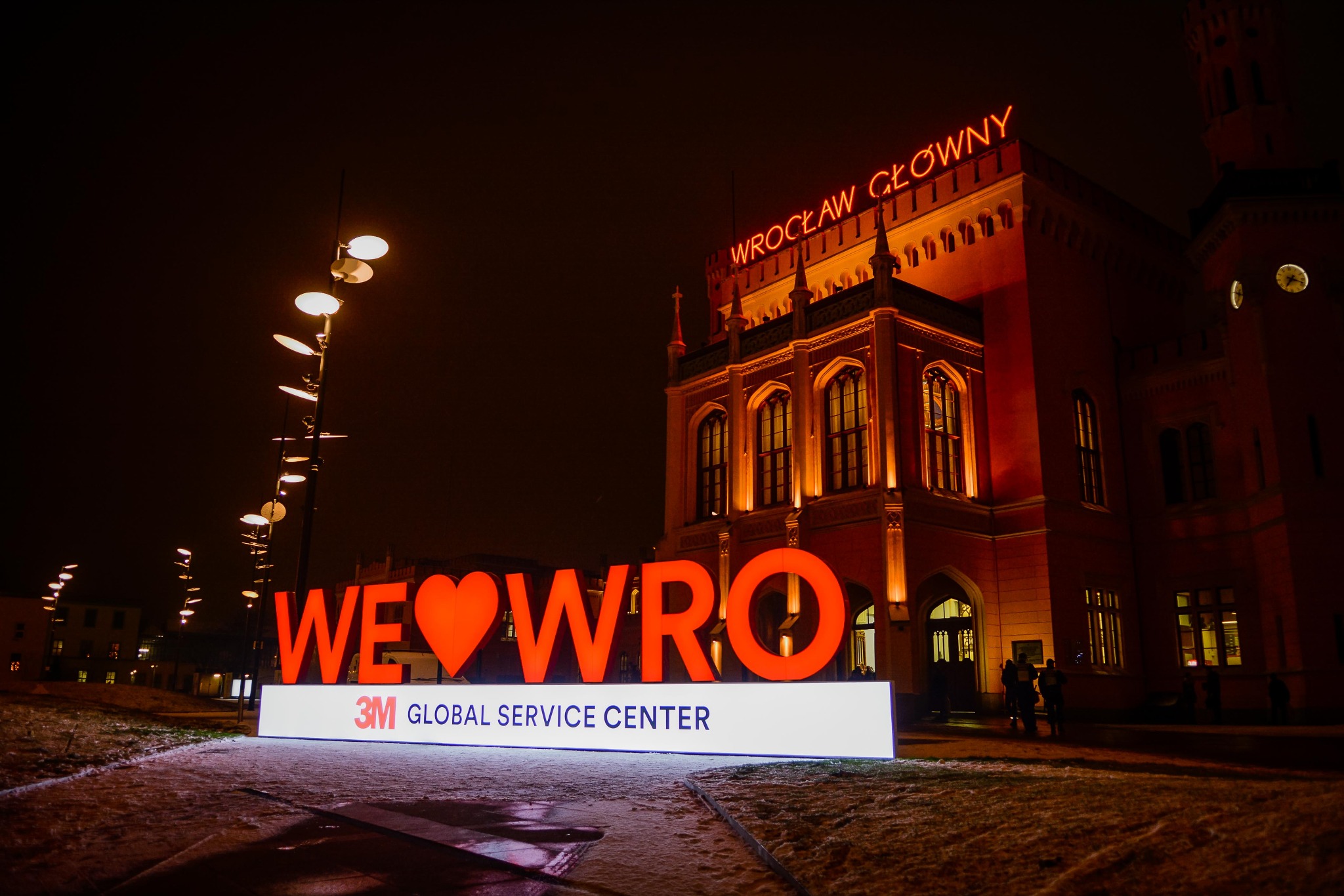 Nocne ujęcie podświetlonej instalacji 'WE❤️WRO' z logo 3M Global Service Center na tle budynku Dworca Głównego we Wrocławiu, oświetlonego ciepłym światłem.