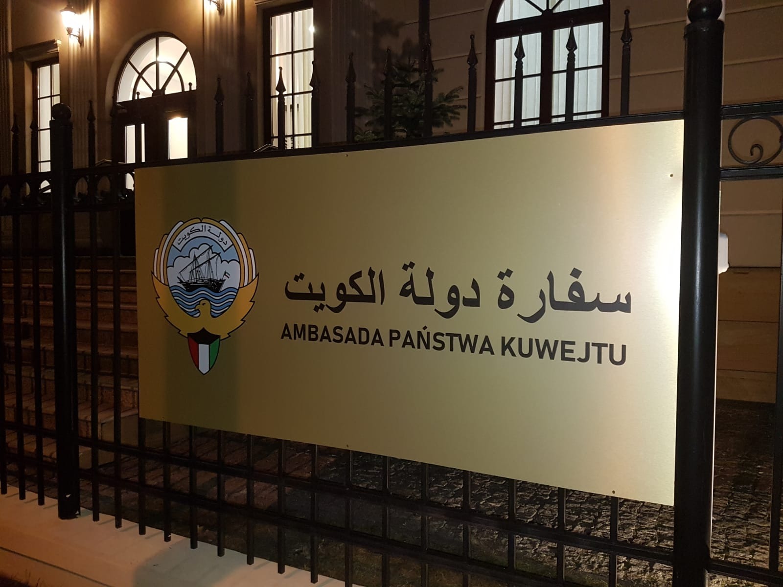 Tablica informacyjna z godłem i napisem 'Ambasada Państwa Kuwejtu' przed budynkiem ambasady, widok z poziomu ulicy.