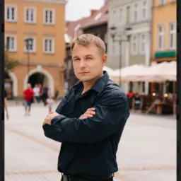 Portret mężczyzny w ciemnej koszuli z założonymi rękami na tle placu miejskiego z kolorowymi kamienicami.