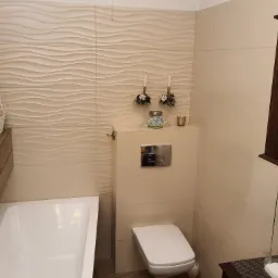 Jasna łazienka z dekoracyjnymi płytkami ściennymi o falującej strukturze, białą wanną i toaletą, oraz wiszącą szafką z ciemnego drewna.