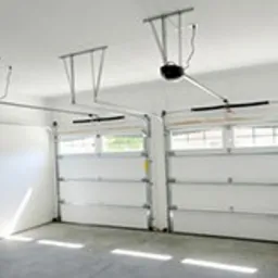 Wnętrze garażu z dwoma białymi, segmentowymi bramami garażowymi, widoczne mechanizmy napędowe pod sufitem, betonowa posadzka z refleksami światła.