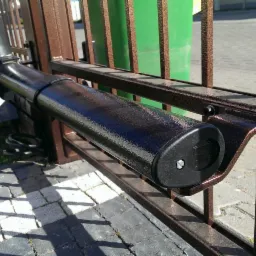 Zamontowany napęd bramy przesuwnej, widoczny mechanizm w czarnej obudowie przymocowany do metalowej, brązowej ramy bramy, z widoczną sprężyną i elementami mocującymi.