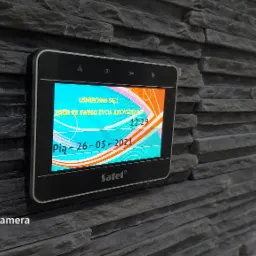 Zamontowany na szarej, kamiennej ścianie panel alarmowy Satel z wyświetlaczem prezentującym datę 26-03-2021, godzinę 12:29 oraz motywacyjne hasło.