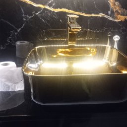 Kris-Rem Remont i Wykończenia wnętrz - Czarna umywalka nablatowa w kształcie prostokąta z pozłacaną armaturą i czarnym marmurem w tle, obok rolka papieru toaletowego.