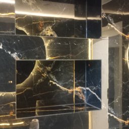 Kris-Rem Remont i Wykończenia wnętrz - Czarne marmurowe płytki z złotymi żyłami tworzą półki we wnęce, podświetlone od góry, z czarnym włącznikiem światła na ścianie i fragmentem armatury.