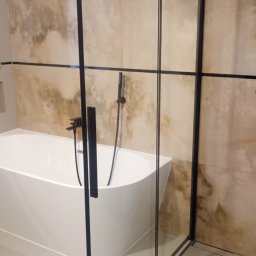 Kris-Rem Remont i Wykończenia wnętrz - Nowoczesna łazienka z wolnostojącą białą wanną i kabiną prysznicową ze szkła z czarnymi profilami, na tle beżowych płytek imitujących marmur.